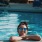 woman with sunglasses, in a pool, rests her arms on the ledge and smiles