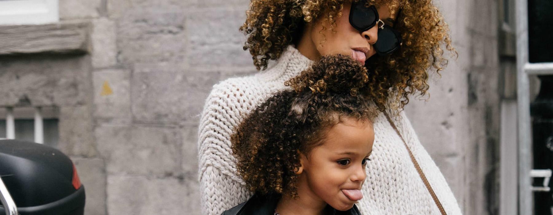 mother and daughter stick their tongues out while looking at themselves in a motorcycle mirror parked in front of a building